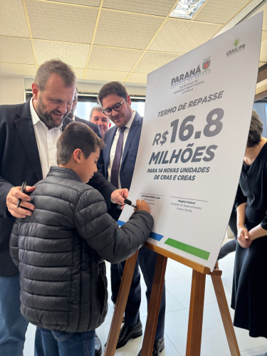Rogério Carboni observa criança assinando placa de repasse de R$ 16,8 milhões para CRAS e CREAS.